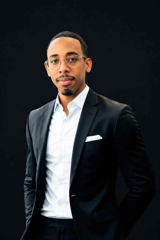 La Jon Dantzler, founder of Dantzler Health Solutions, photographed from the waist up in a black suit jacket, white open-collar shirt and glasses with a gold frame, against a dark studio background
